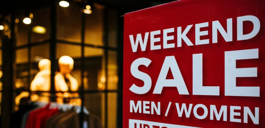  A red sign in a clothing store window advertising a "Weekend Sale" for men's and women's items, offering discounts up to a certain percentage. The background shows mannequins and clothing displays inside the store.