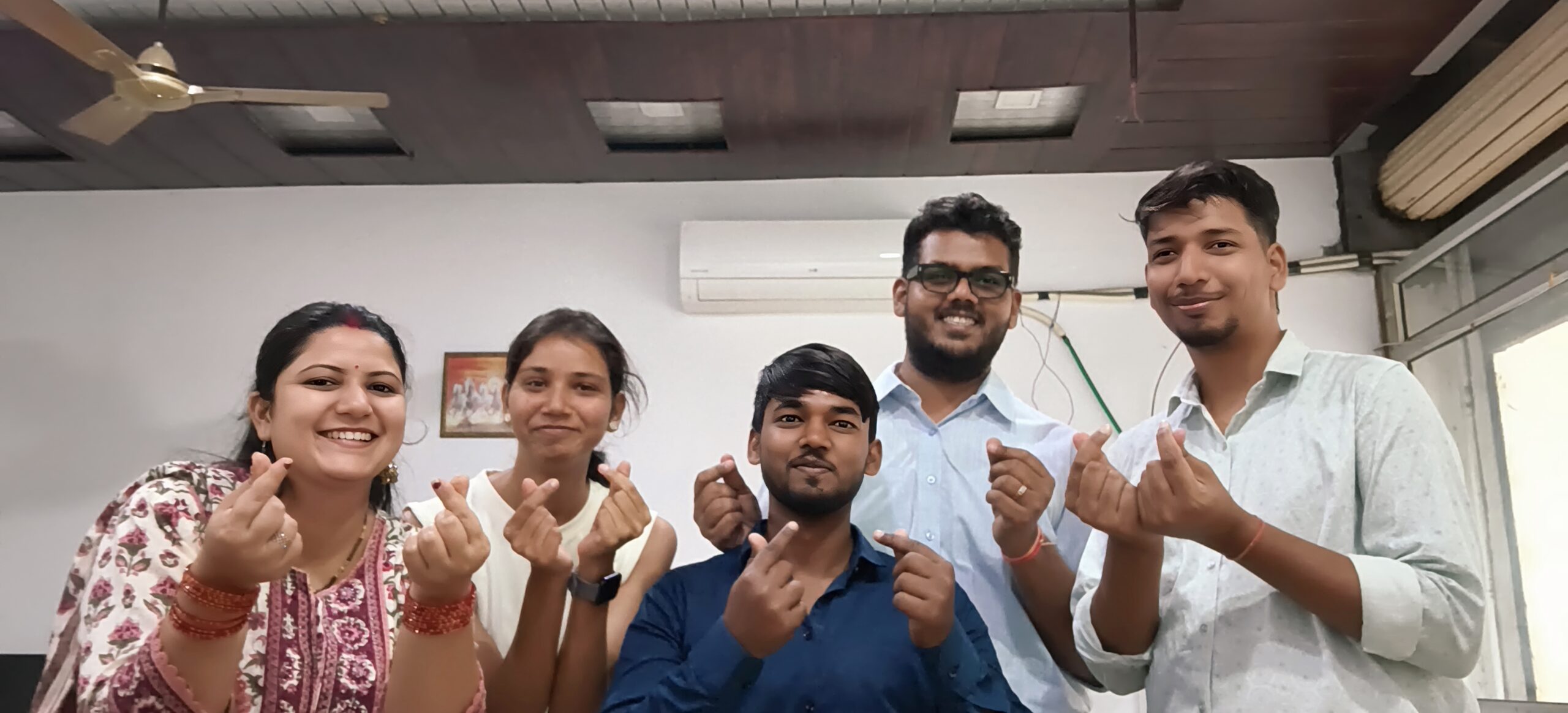 A group of six colleagues smiling and making finger heart gestures inside an office, capturing a warm and emotional farewell moment at a digital marketing workplace.